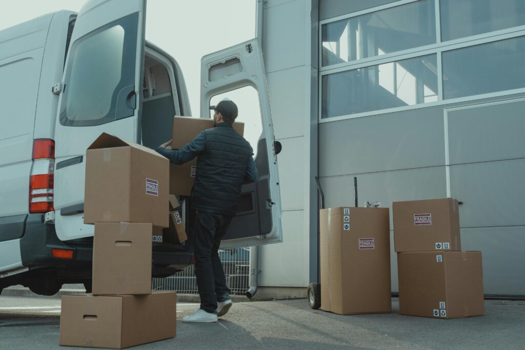 A delivery man unloading cardboard boxes from a van at a warehouse during the day.